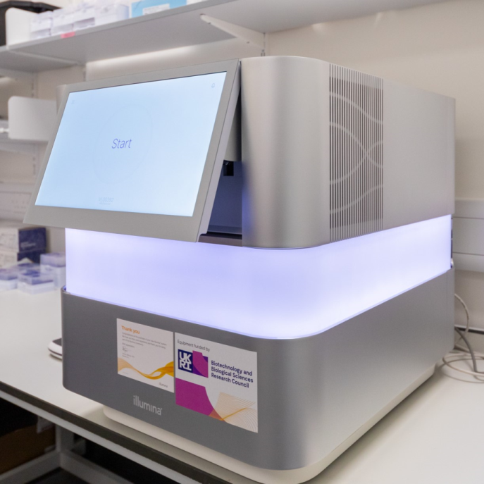 Image of the NextSeq 1000 sequencer. This sequencer is a large silver cube with a wide strip of LED lighting around the centre of the instrument, currently display white and purple colouring. The instrument has a tablet on the front of it, displaying a white screen and the word 'start'. 